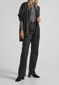 Camisa de cuero negro superpuesta a una blusa de manga larga con estampado púrpura, combinada con pantalones oscuros de cintura alta y sandalias de tacón negras.