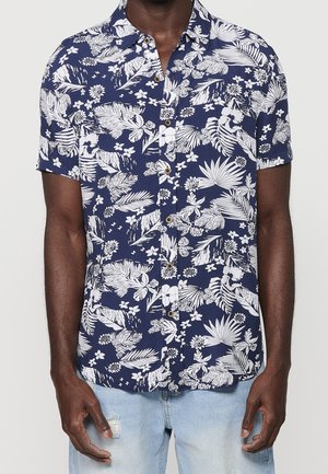 Chemise bleu marine à manches courtes avec boutons, motif floral et feuilles tropicales blanches, portée avec un jean bleu clair usé.