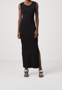 Robe maxi noire sans manches avec une fente latérale, texture douce, coupe près du corps, associée à des sandales argentées brillantes à boucles.