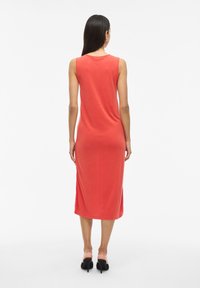 Vestido midi sin mangas de color coral con cuello redondo, textura suave y corte relajado. Presenta una abertura lateral en el dobladillo y se combina con tacones negros.