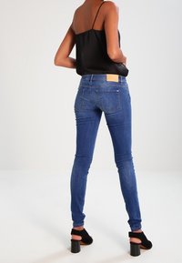 Jeans ajustados azul con una textura de denim suave, tiro medio, y una etiqueta de cuero marrón claro en la parte trasera. Sin adornos visibles.