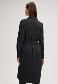 Zwarte, langslijvende jurk met een kraag, ceintuurlijn en achterkant met plooistiksel. De stof oogt glad met een matte afwerking. Knopen op de manchetten.