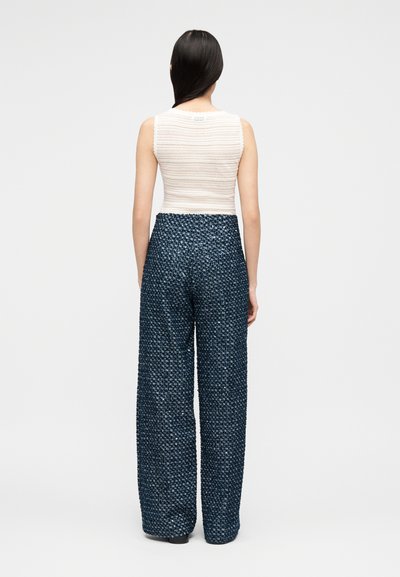 Γυναίκα με μακριά σκούρα μαλλιά που φοράει λευκό αμάνικο τοπ με υφή και φαρδιές ψηλόμεσες μπλε σκούρες παντελόνες, όρθια με την πλάτη γυρισμένη.