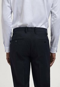 Navy dress pants with a straight cut, flat front, two back pockets with buttons, worn with a light blue striped dress shirt.