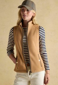 Beige Fleece-Weste mit Reißverschluss vorne und seitlichen Taschen, getragen über einem navy-blauen und weißen gestreiften Langarmshirt. Mit goldenen Akzenten.