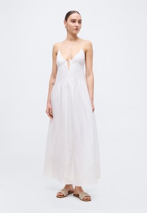 Kvinde iført en hvid maxi-kjole uden ærmer med tynde stropper og dyb V-udskæring, kombineret med beige vævede slid-sandaler.