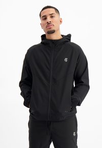 Zwarte lichtgewicht zip-up jack met capuchon, voorzien van een subtiele logo, elastische manchetten en een ontspannen pasvorm. Soepele textuur en minimaal ontwerp.