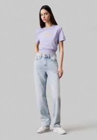 Camiseta de algodón lila con texto amarillo, combinada con jeans de pierna recta en azul claro. El modelo lleva zapatillas blancas. Fondo gris neutro.