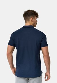 Hombre con cabello corto y rizado, vestido con una camisa polo azul marino de manga corta, de espaldas contra un fondo liso gris claro.