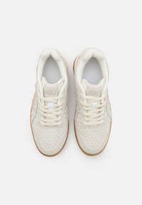 Scarpe sportive beige con tomaia in pelle, punta forata, lacci piatti e suola in gomma. Branding visibile sulla linguetta.
