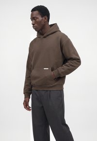Homme avec la main dans la poche portant un sweat à capuche marron et un pantalon gris foncé, debout devant un fond clair uni.