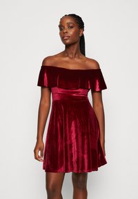Burgundské off-shoulder šaty z jemného zamatového materiálu. Obsahujú priliehavú korzetovú časť, rozšírenú sukňu a volánikový výstrih. Hladká textúra po celej ploche.