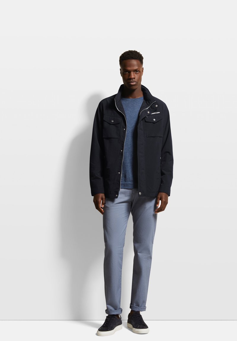 Schwarze Jacke mit zwei Fronttaschen, getragen über einem blauen Pullover, kombiniert mit hellgrauen Hosen und dunklen Schuhen. Schlichtes, modernes Design.