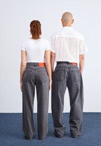Jean large gris avec des étiquettes orange à la taille, portés par deux personnes de dos. L'une porte un t-shirt blanc court, l'autre un haut à motifs.