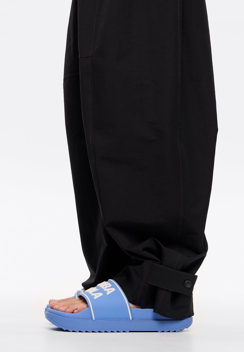 Sandali blu con testo bianco, suola testurizzata e una fascia larga, abbinati a pantaloni neri larghi con cuciture arancioni e dettagli con bottoni.