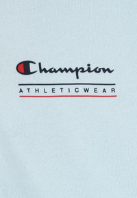 Gaiši zils sporta t-krekls, kas izgatavots no teksturēta auduma, ar melnu "Champion" logo un sarkanu akcentu, zem kura ir teksts "ATHLETICWEAR".