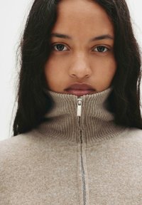 Lynlås sweater i lys beige med ribbet krave og subtil tekstur, samt en metallisk lynlås fortil.