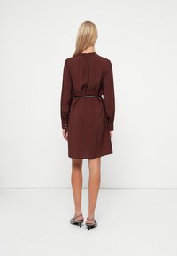 Καφέ φραμπαλά φορεματάκι από ελαφρύ ύφασμα, με μακριά μανίκια, κουμπωτές μανσέτες και άνετη εφαρμογή. Η γραμμή του τελειώνει στο μέσο του μηρού.