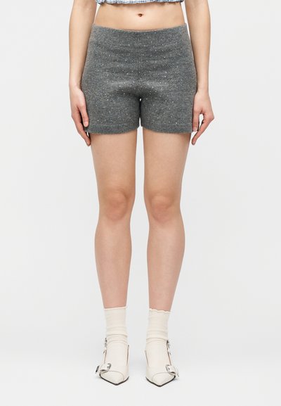 Γυναίκα που φοράει γκρι πλεκτές σορτς με διακριτικό πουά σχέδιο, λευκές κάλτσες και λευκά μυτερά παπούτσια με διακοσμητικές αγκράφες, στέκεται μπροστά σε απλό φόντο.