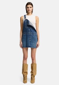 Abito da salopette in denim con parte superiore chiara e tasche frontali, abbinato a stivali alti marroni, texturizzati e con accenti metallici.
