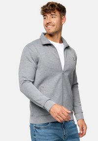 Graue Kapuzenjacke aus weichem Material, mit hohem Kragen, langen Ärmeln und seitlichen Taschen, kombiniert mit blauen Jeans.