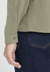 Olivgrünes Langarmhemd mit einem Knopf am Ärmelsaum, getragen mit dunkelblauen Jeans. Sichtbare Nähte und lockere Passform.