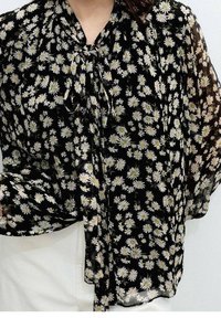 Blouse noire à motifs floraux avec marguerites blanches, tissu léger, coupe ample et col noué. Met en valeur une texture fluide et transparente.