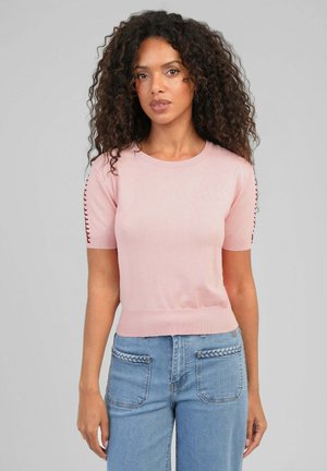 Femme aux cheveux bouclés portant un pull rose à manches courtes avec des détails de couture sur les manches et un jean bleu clair avec une bordure tressée aux poches.