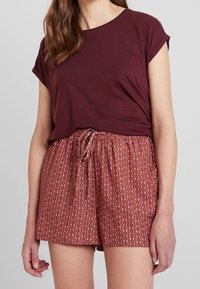 Burgundy topp med uppvaktade ärmar, parat med mönstrade röda shorts med elastisk midja och dragsko. Geometrisk design synlig.