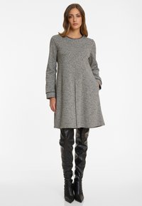 Frau in einem grauen, strukturierten Langarmkleid mit schwarzem Saum und schwarzen, obsidian hohen Lederstiefeln, stehend vor einem weißen Hintergrund.