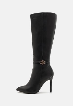 Botte en cuir noire haute jusqu'au genou, avec bout pointu, talon stiletto et fine lanière décorative ornée d'un emblème rond doré autour de la cheville.