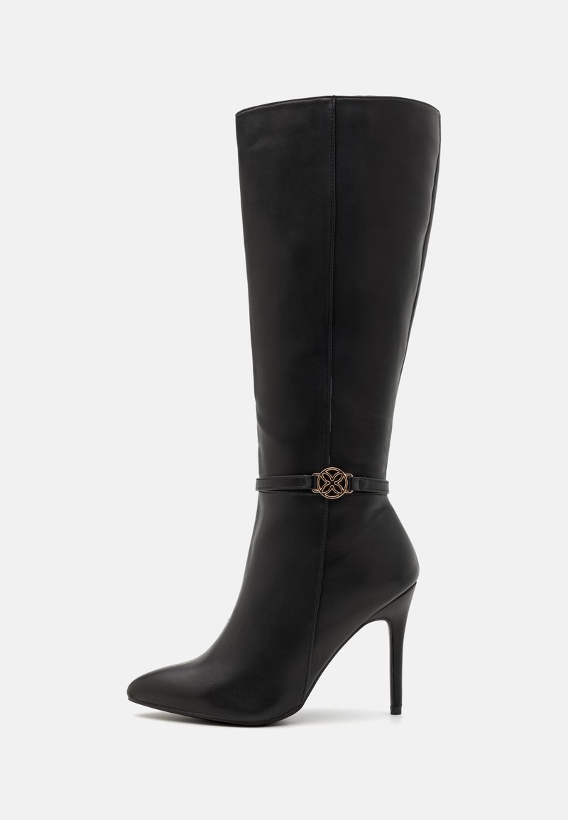 Botte en cuir noire haute jusqu'au genou, avec bout pointu, talon stiletto et fine lanière décorative ornée d'un emblème rond doré autour de la cheville.