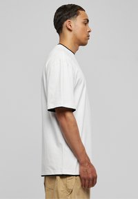Vit bomull t-shirt med korta ärmar och svart kontrasterande kant längs halsringningen och ärmsluten, sedd från sidan.