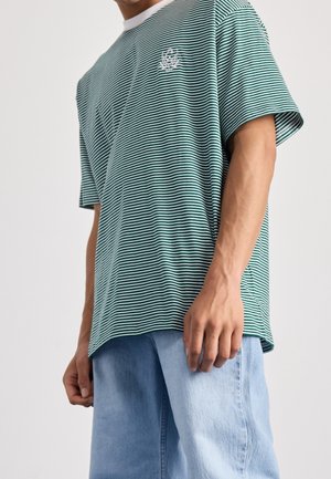 Person mit einem lockeren grün-weiß gestreiften T-Shirt mit kurzen Ärmeln und hellblauen Jeansshorts steht vor einem schlichten Hintergrund.