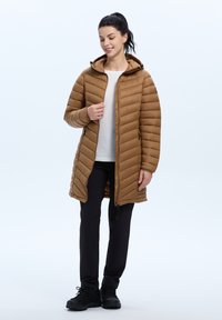 Bruine gewatteerde pufferjas met een capuchon, voorzien van verticale stiksels, een rits aan de voorkant en twee zijzakken, gedragen over een wit topje en zwarte broek.