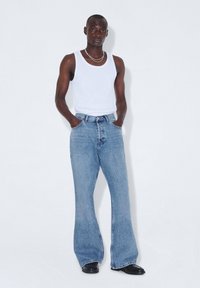 Jeans de mezclilla azul de pierna acampanada con cintura alta y un lavado claro, combinados con una camiseta sin mangas blanca. Los zapatos son negros con un acabado liso.