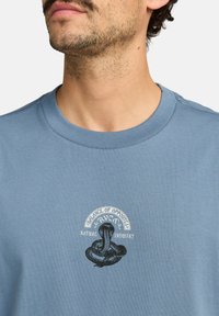 Camiseta de algodón azul con cuello redondo, que presenta un gráfico negro de una cobra y el texto: "BALANCE DE OPUESTOS RVCA INDUSTRIA NATURAL."