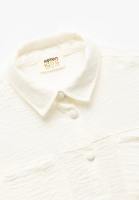 Camicia per bambini in cotone bianco con colletto, chiusura frontale a bottoni e due tasche sul petto, dotata di una superficie testurizzata e bottoni bianchi rotondi.