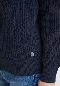 Pull en tricot bleu marine avec une texture côtelée, détail du logo en argent rond sur l'ourlet et poignets ajustés. Porté avec un jean bleu clair.