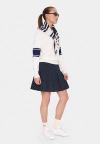 White sweater with navy stripes, navy pleated skirt, and white sneakers. A striped scarf is tied at the neck, adding detail.