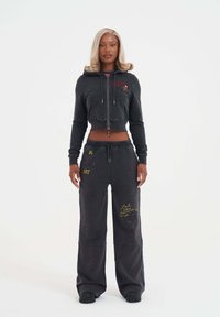 Avskuren svart hoodie med framfickor och broderade detaljer, kombinerad med lösa svarta byxor med gula textdetaljer. Tillverkad av mjukt tyg.