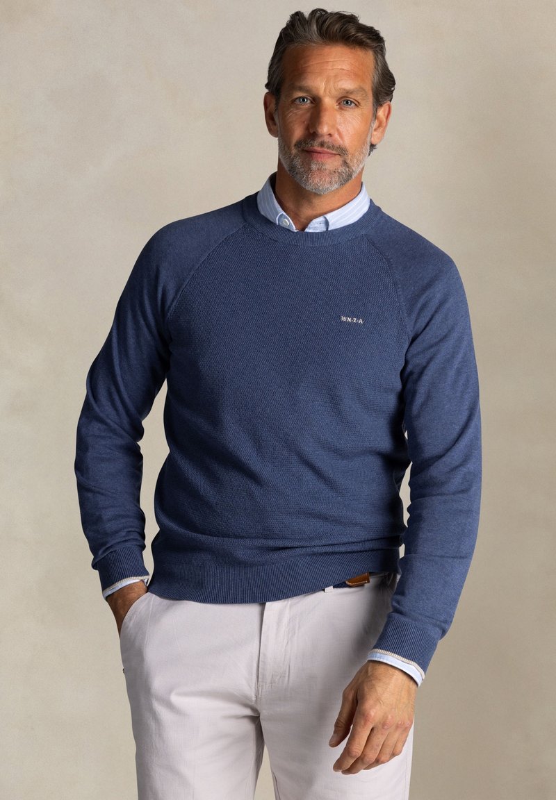 Aansluitende blauwe crewneck trui van textuurstof, met ribgebreide manchetten en zoom, gedragen over een lichtblauw overhemd met kraag, gecombineerd met lichte broek.