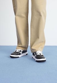 Des baskets noires avec des semelles et des lacets blancs, présentant un design supérieur texturé. Portées avec un pantalon beige clair sur une surface bleue.