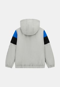 Svijetlosiva jakna s kapuljačom koja ima plave i crne akcentne detalje na rukavima, opuštenog kroja i elastične manžete na zapešćima.