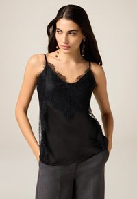 Schwarzes Camisole mit Spitzenbesatz rund um den Ausschnitt und an den Seiten, mit dünnen Trägern. Der Stoff ist durchsichtig und hat eine glatte Textur.