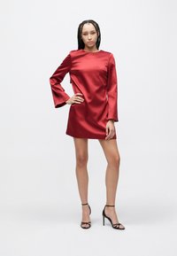 Kvinne med fletter iført en rød minikjole i sateng med lange ermer og svarte høyhælte sandaler som står foran en ensfarget bakgrunn.