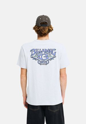 Persoon draagt een wit Billabong t-shirt met een blauw logo en tekst op de achterkant en een donkere pet, met de rug naar de camera gekeerd.