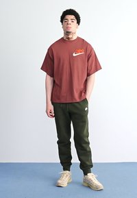 Tumši sarkans Nike T-krekls ar baltu logo, savienots ar tumši zaļiem treniņbiksēm un bēšiem sporta apaviem. Istaba ar gaišu fonu un zilu grīdu.