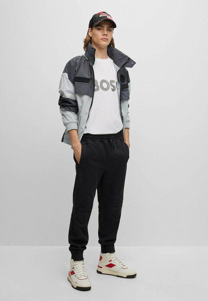 Giovane con un cappellino nero, maglietta con logo bianco, giacca grigia e nera, pantaloni neri e sneakers bianche con dettagli rossi, che sta in piedi con le mani in tasca.