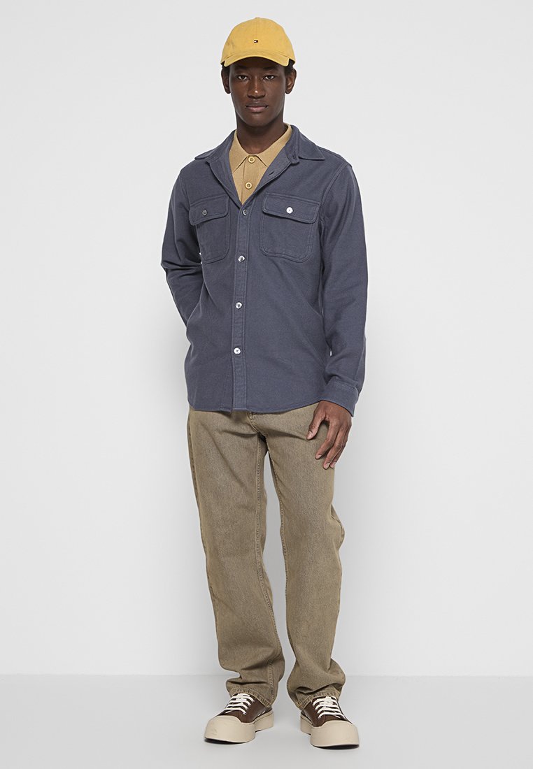 Mann, der eine gelbe Kappe, ein hellbraunes Polohemd, eine blaue knopffrontige Jacke, beigefarbene Hosen und braun-weiße Turnschuhe trägt, steht vor einem weißen Hintergrund.